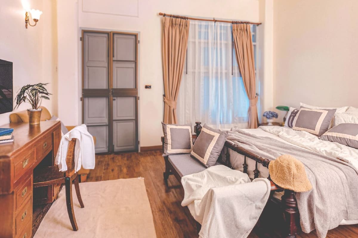 A bedroom with a wooden bed, a chair, a desk, a potted plant, and a window with curtains.