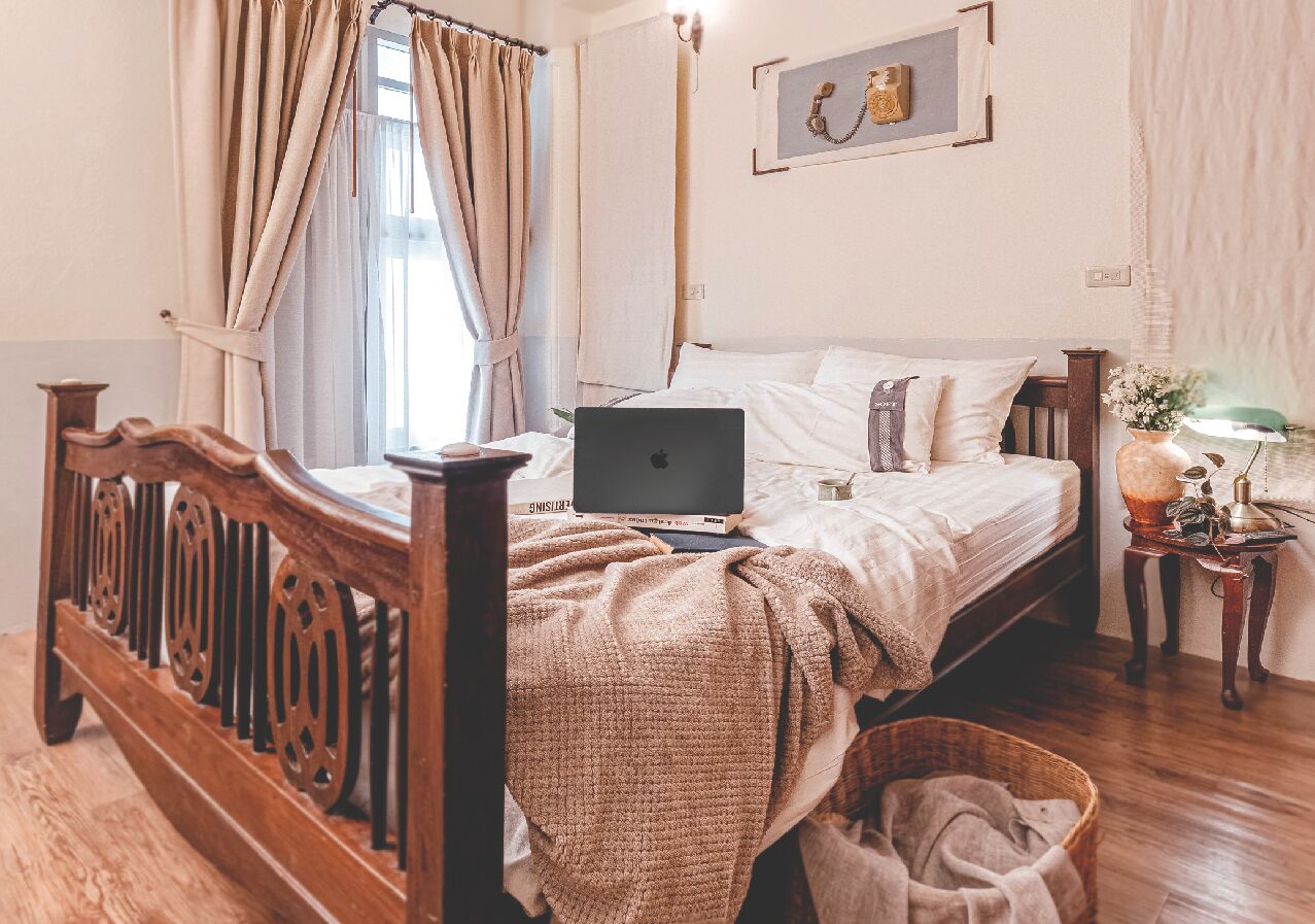 A bedroom with a wooden bed, a laptop on the bed, a bedside table with a lamp, and a window with curtains.