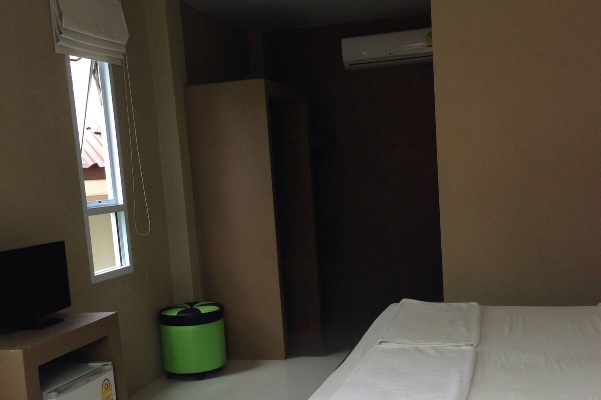 Una habitación de hotel con cama, televisión, refrigerador pequeño, basurero y ventana con persianas.