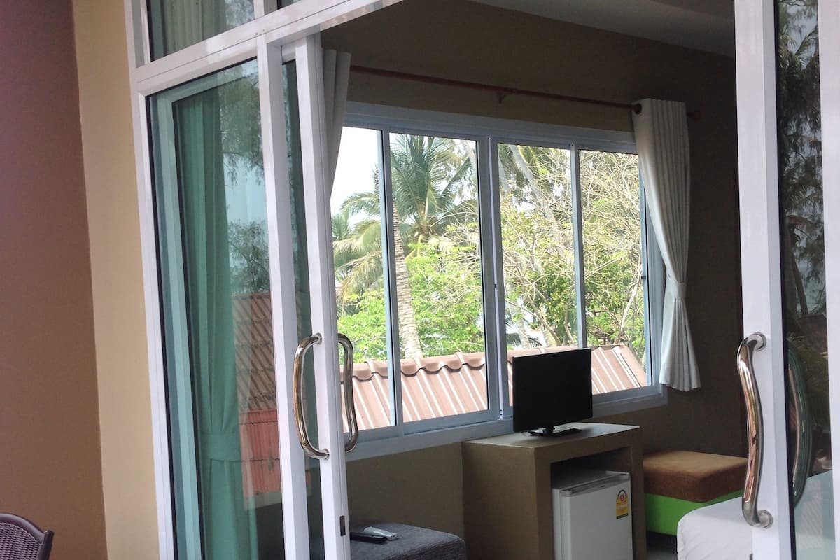 Una habitación con puerta de vidrio corredera que da a un balcón, un escritorio pequeño con una computadora, una zona de estar con cojines y una silla.