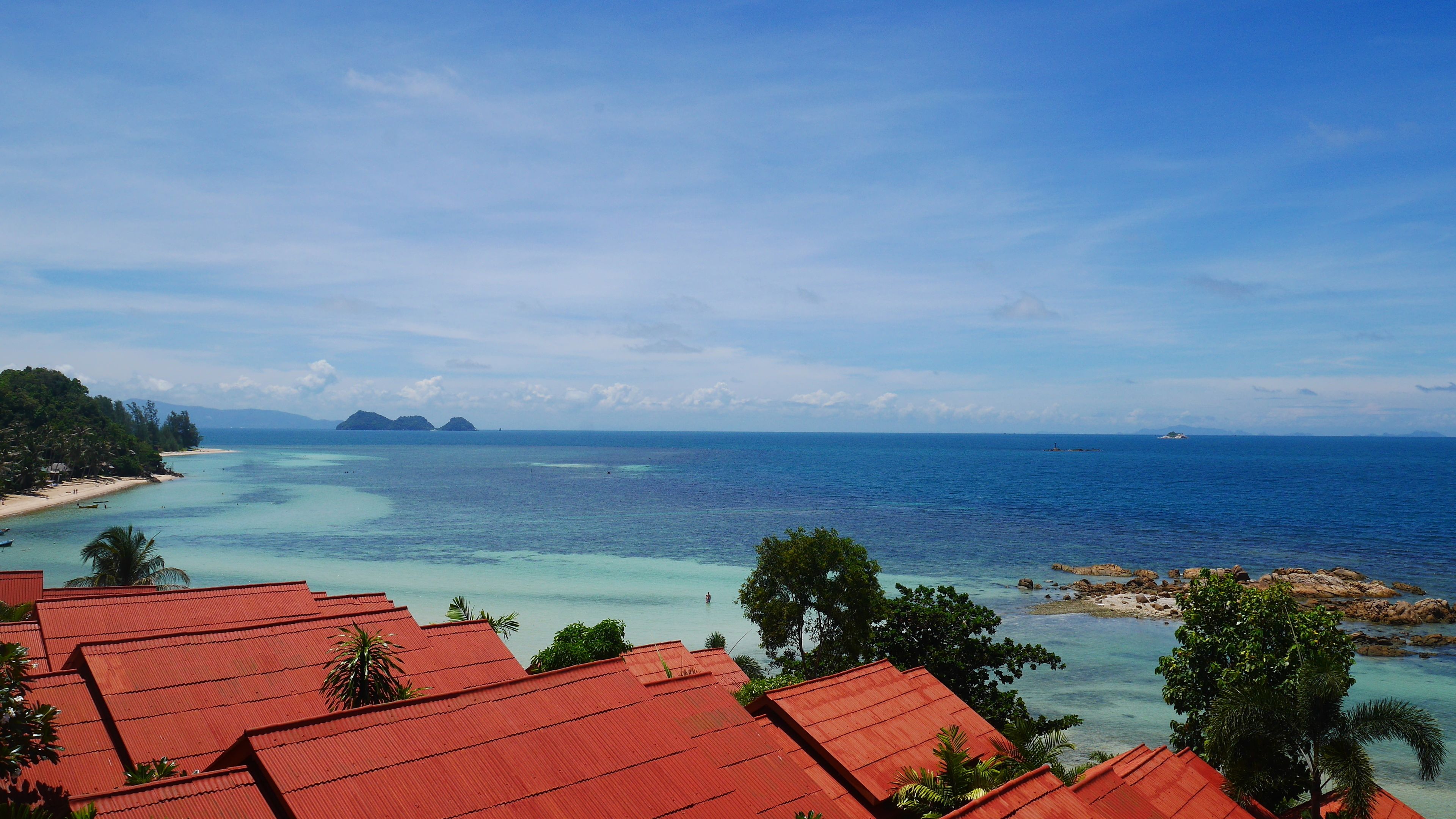 A coastal view with clear turquoise waters, distant islands, and a red-roofed building in the foreground.