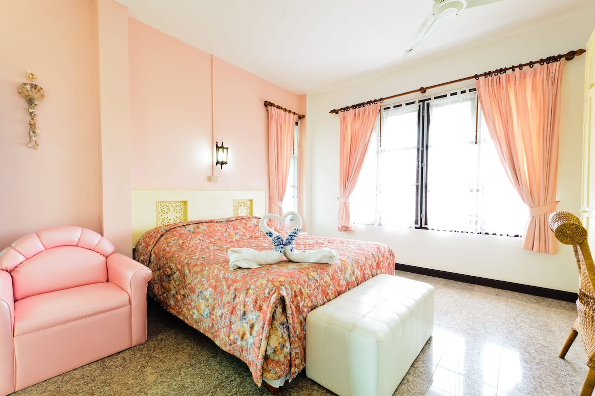 Une chambre à coucher avec une couvre-lit à motifs floraux, une chaise rose, un banc blanc et un élément décoratif mural.