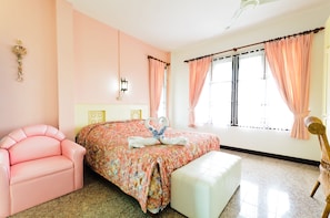 Quarto com colcha florida, uma cadeira rosa, um banco branco e um objeto decorativo na parede.