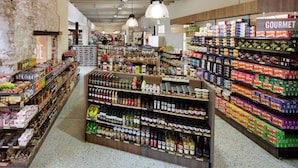A grocery store interior with shelves stocked with various products, a 'GOURMET' section, and a rustic stone wall on the left. - Stay Rottnest (Rottnest Island)