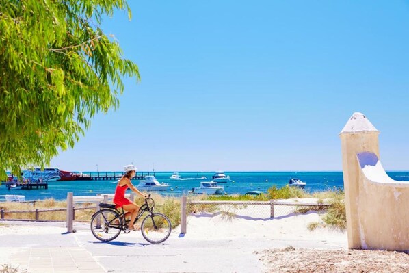 A coastal scene with a person riding a bicycle on a paved path, overlooking a marina with boats.