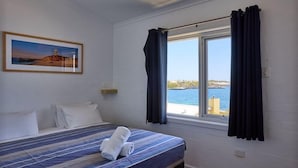 A hotel room with a bed, a painting of a lighthouse, a window with curtains, and a view of the sea. - Stay Rottnest (Rottnest Island)