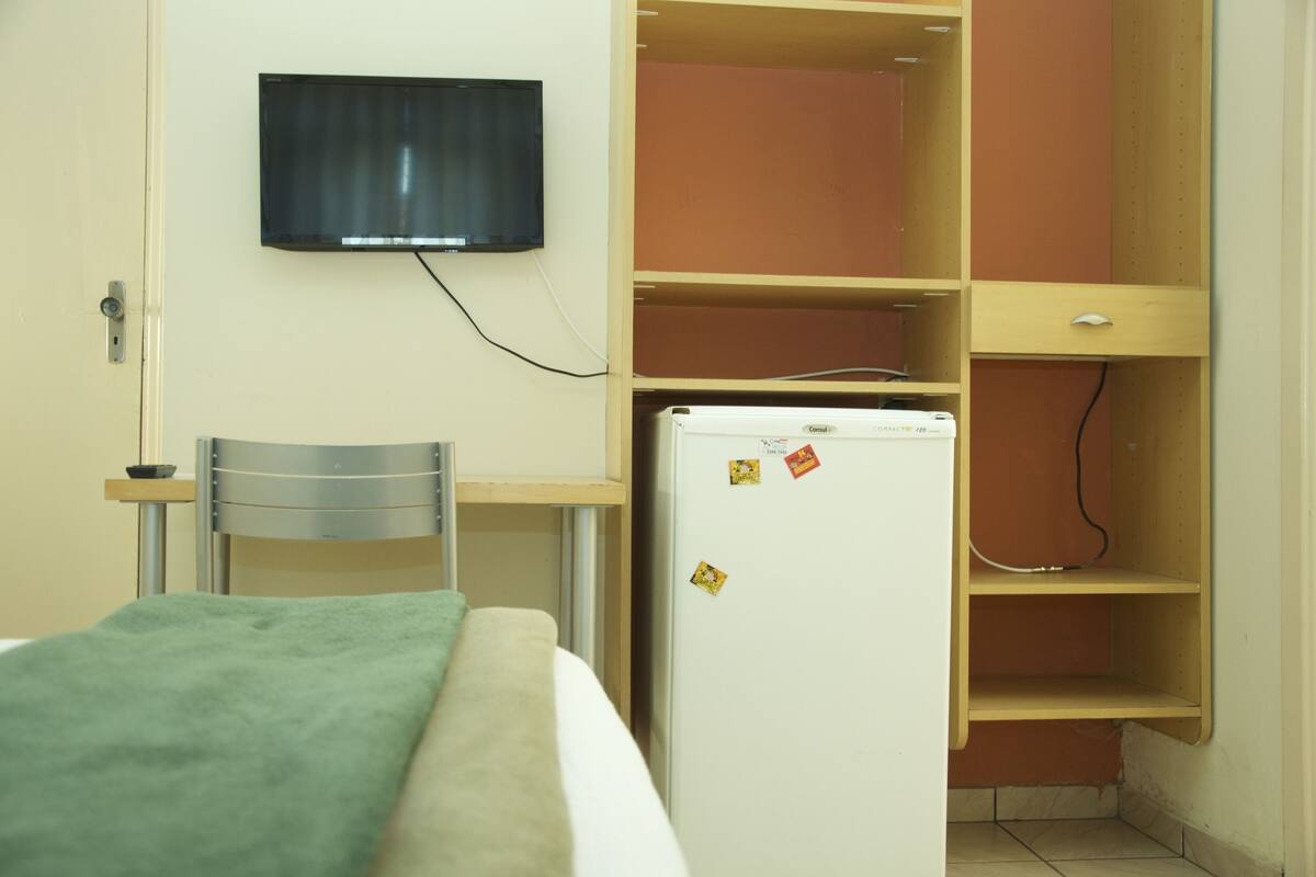 A small room with a bed, a desk, a chair, a television mounted on the wall, and a refrigerator.