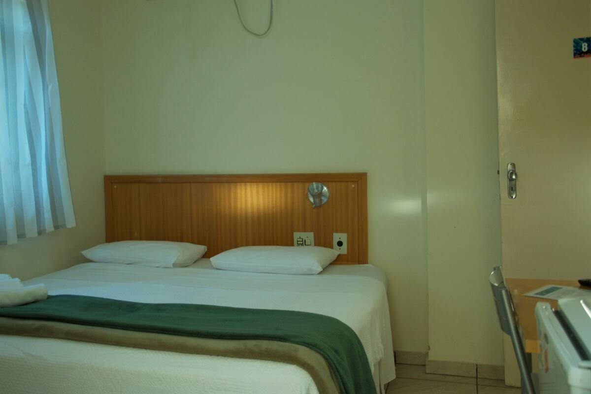 A hotel room with a bed, a wooden headboard, a window with curtains, and a door with a handle.