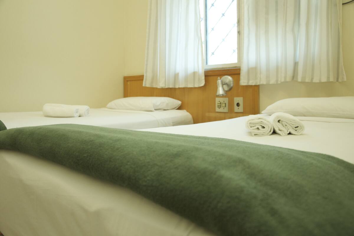 A hotel room with two beds, a green blanket, white towels, a wooden headboard, and a window with curtains.