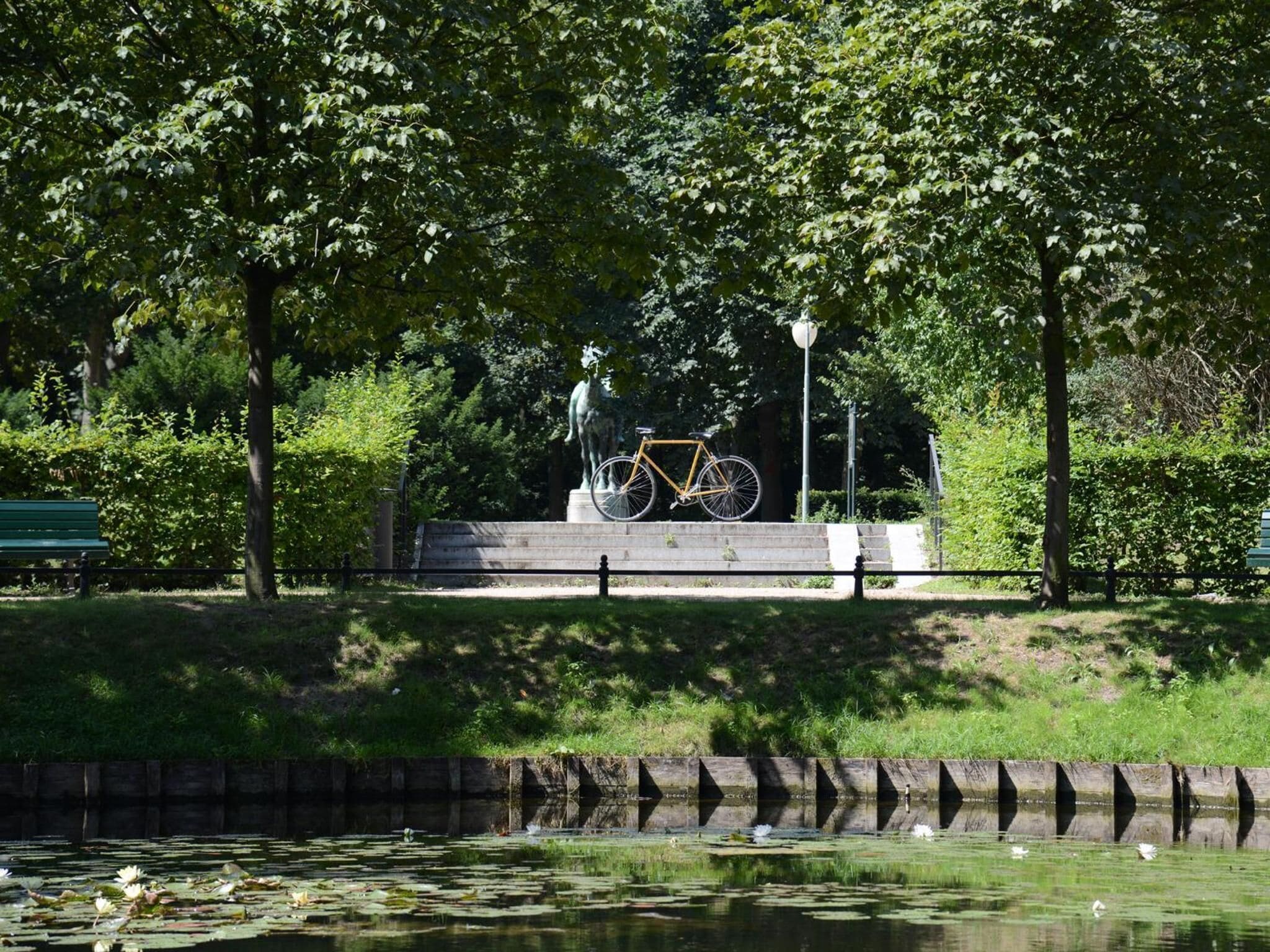 A park with a statue of a person on a bicycle, a bench, and a pond with water lilies.