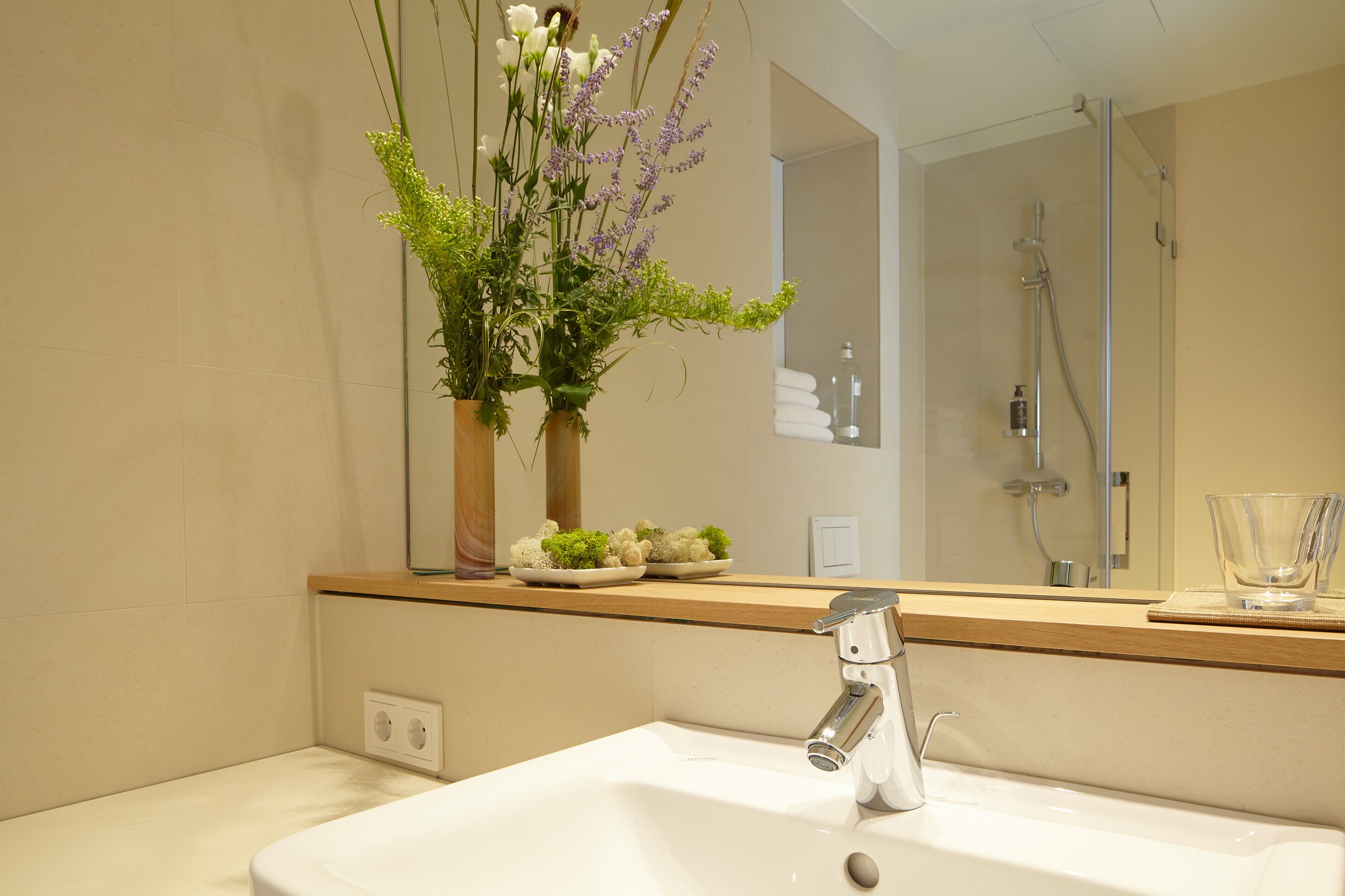 A bathroom with a white sink, a mirror, and a shower. There are two vases with flowers on the counter.