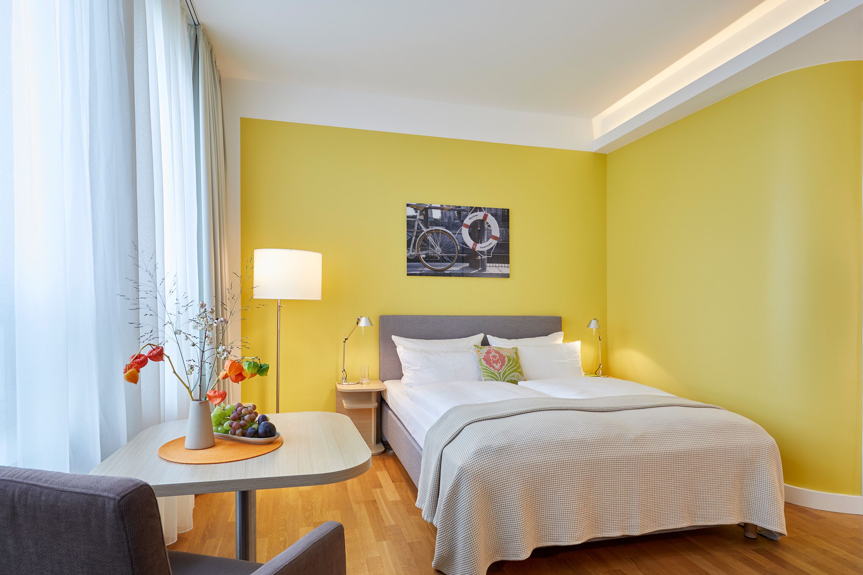 Una habitación de hotel con cama, mesitas de noche, lámpara, una pequeña mesa con un florero y un cuadro en la pared.