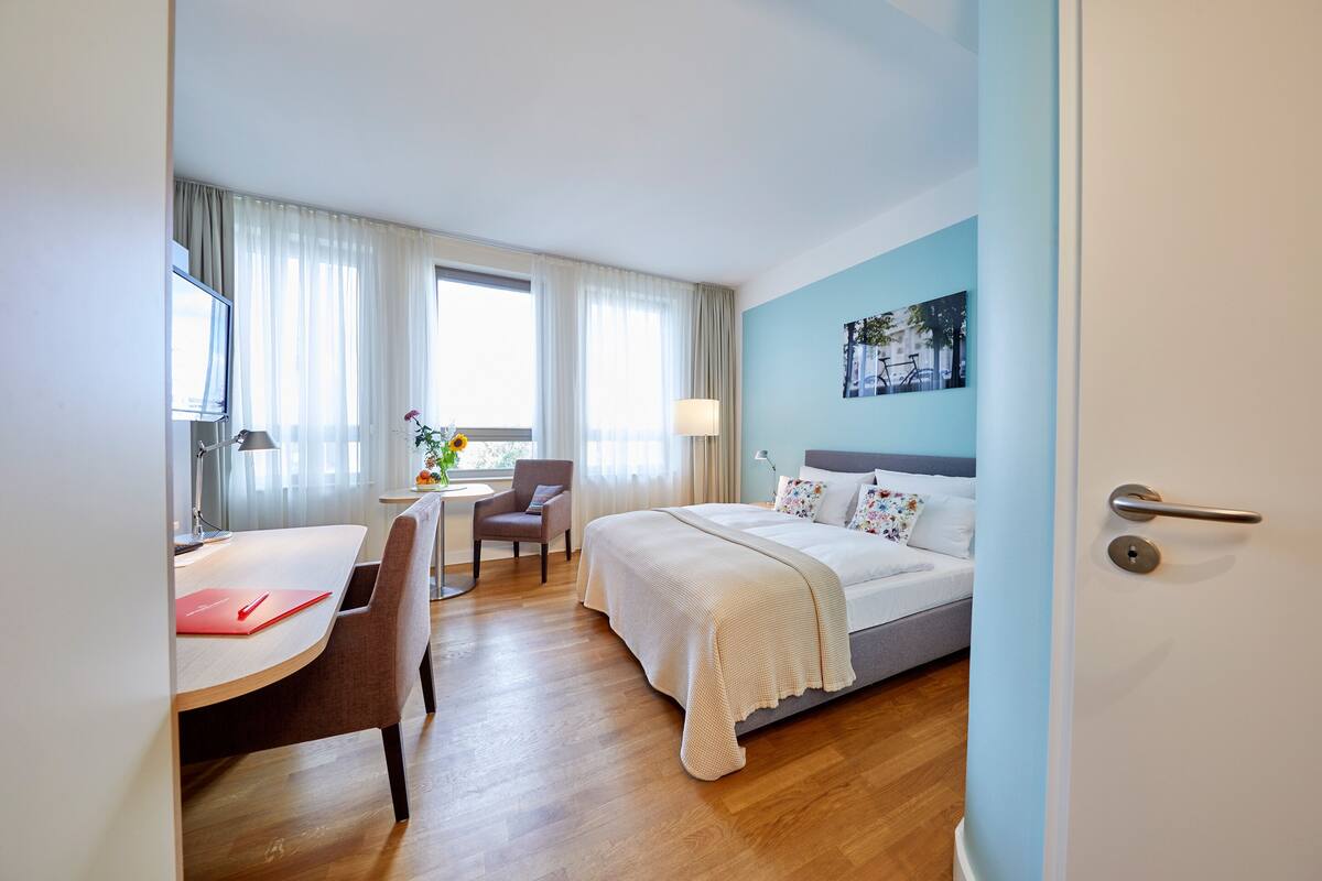 A hotel room with a bed, a desk, a chair, and a window with curtains.