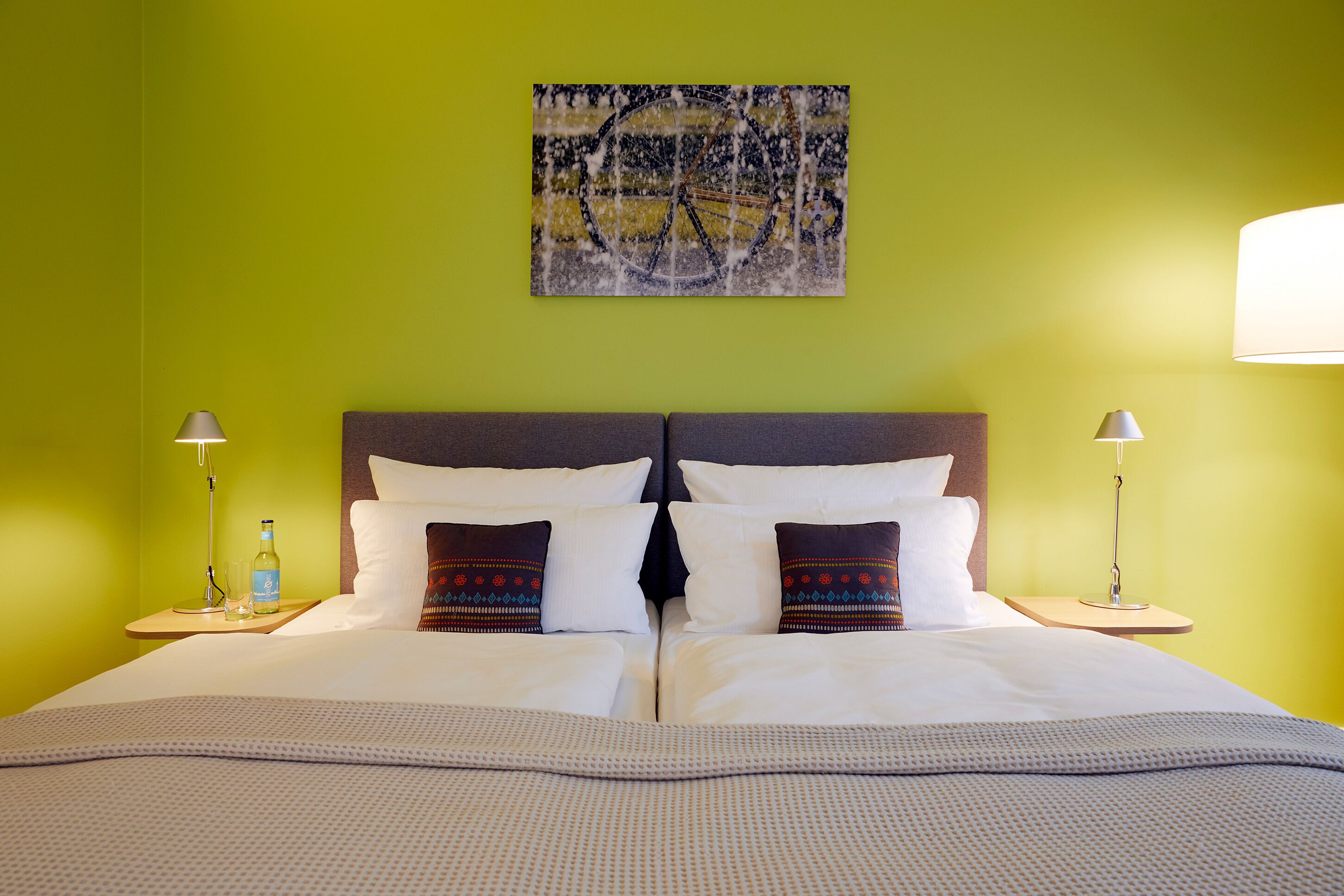Cama bem arrumada com roupa de cama branca, duas almofadas decorativas, uma cabeceira e uma obra de arte na parede.
