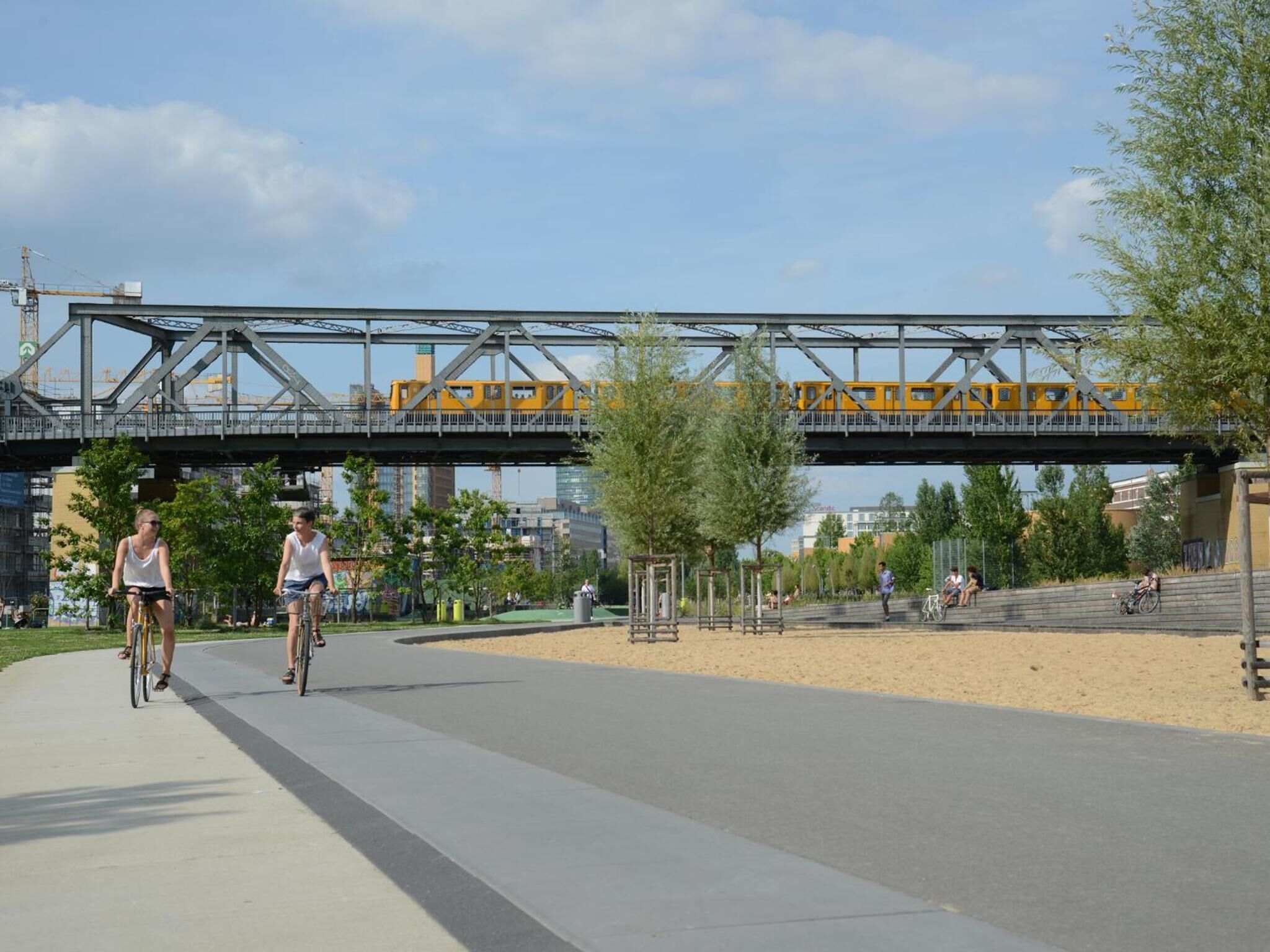 A paved pathway with cyclists, a bridge in the background, and a sandy area with trees.