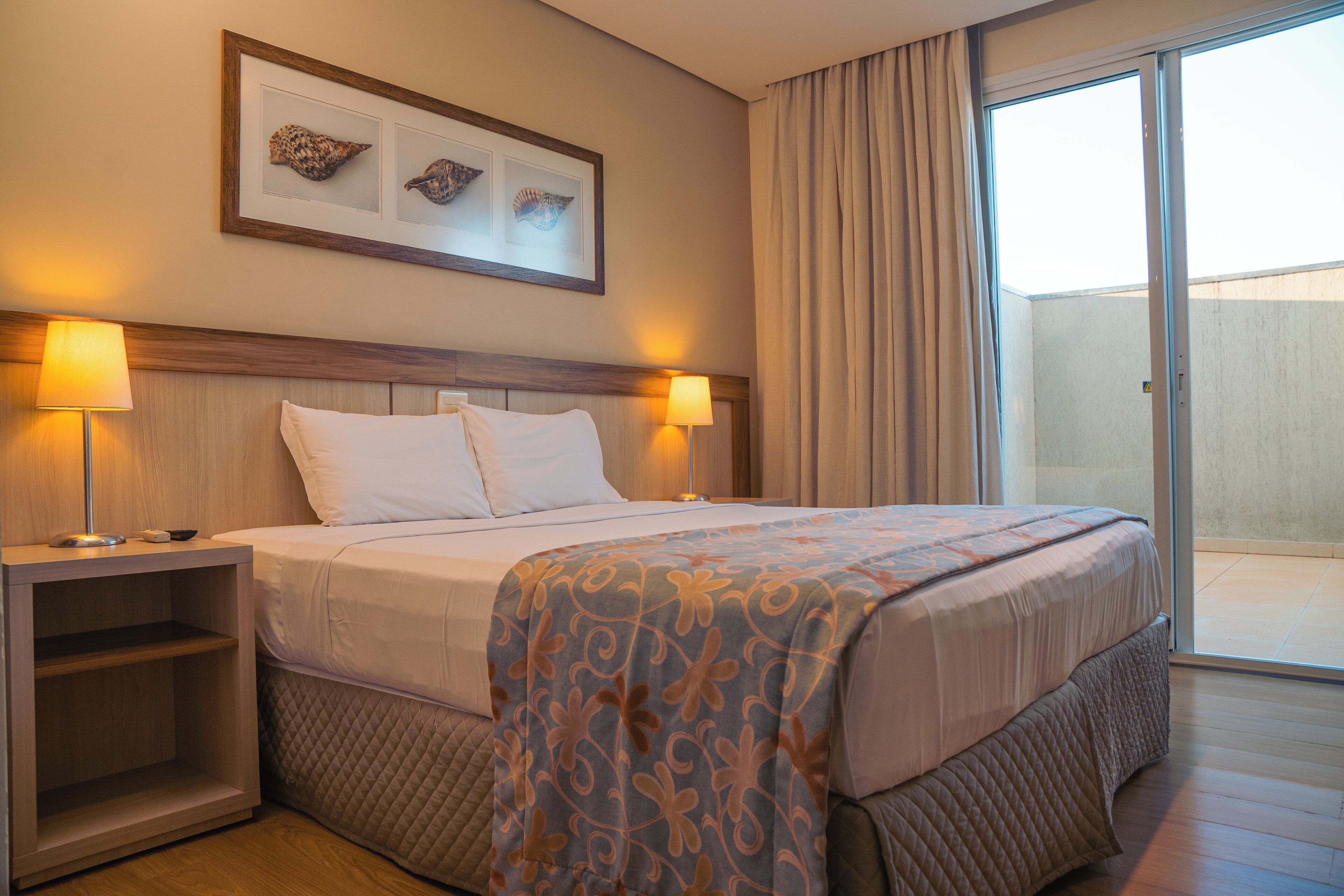 Una habitación de hotel con una cama, dos lámparas de noche, una mesita de noche, un ventanal grande y un cuadro en la pared.