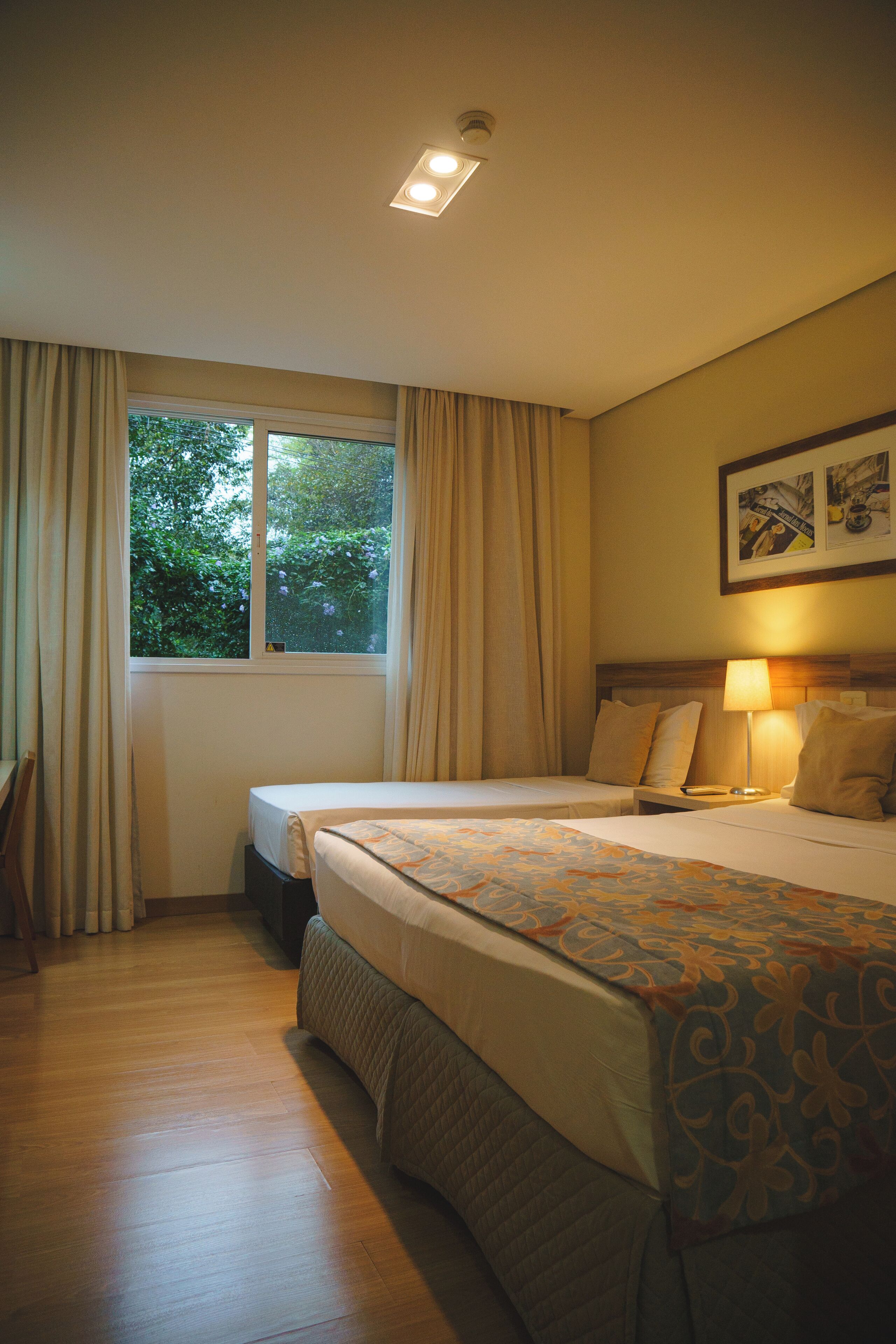Ein Hotelzimmer mit zwei Betten, einem Fenster mit Vorhängen, einem Nachttisch mit Lampe und einem gerahmten Bild an der Wand.