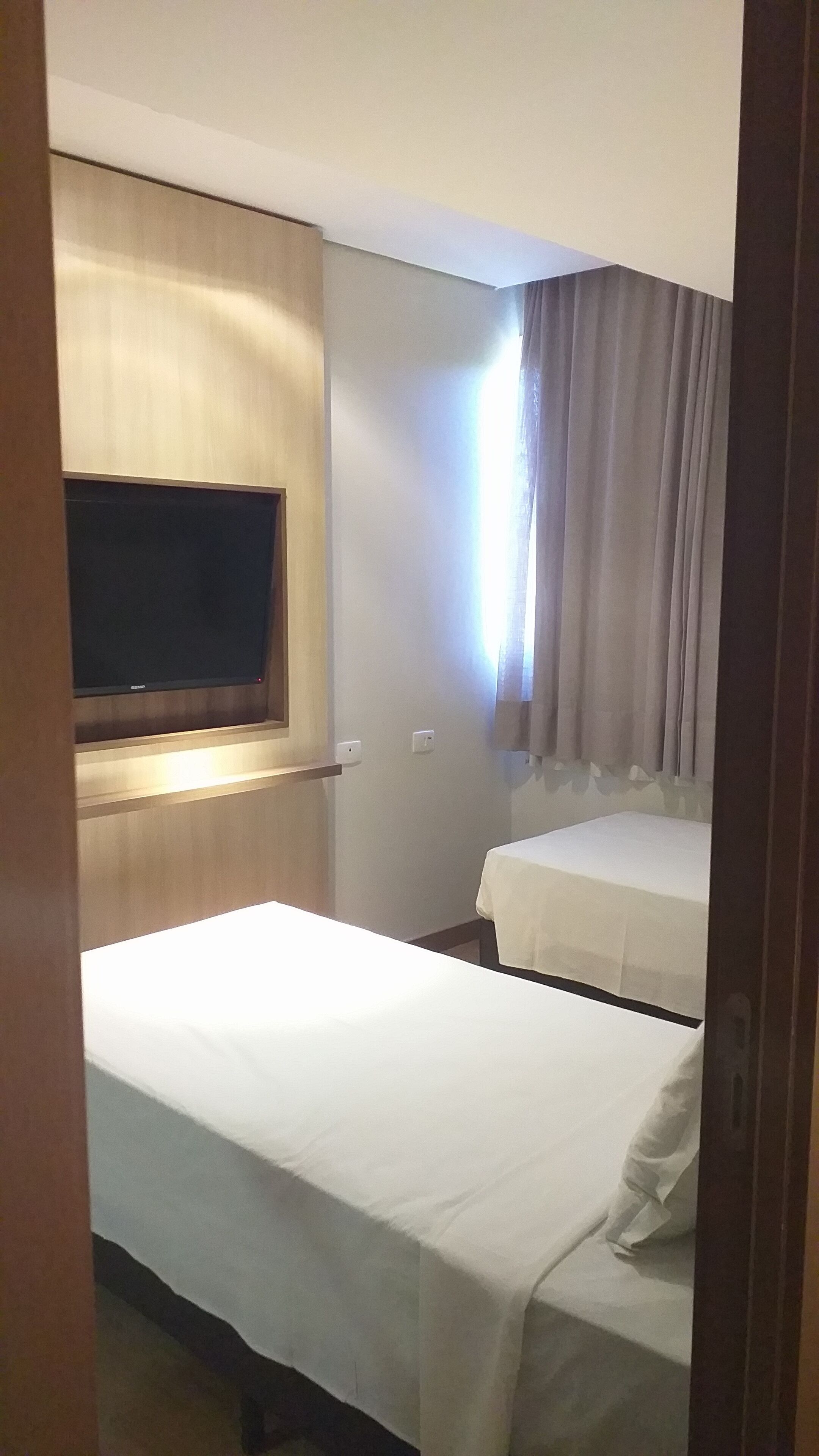 A hotel room with a bed, a TV mounted on the wall, a window with curtains, and a bedside table.