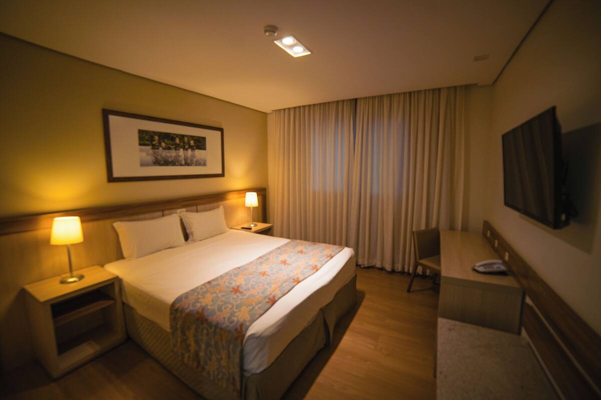 Een hotelkamer met een bed, nachtkastjes, een bureau, een stoel, een televisie en een raam met gordijnen.