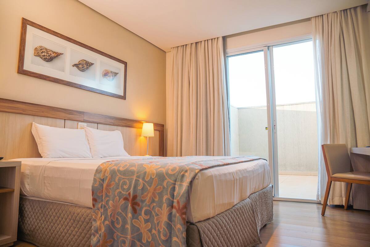 Een hotelkamer met een bed, nachtkastje, stoel, bureau, raam met gordijnen en een wanddecoratie.