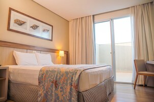 Una habitación de hotel con cama, mesita de noche, silla, escritorio, ventana con cortinas y cuadros en la pared.