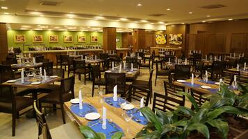 A restaurant with multiple dining tables set with blue placemats, white napkins, and candles. There are green walls with framed pictures and a wooden bar area.
