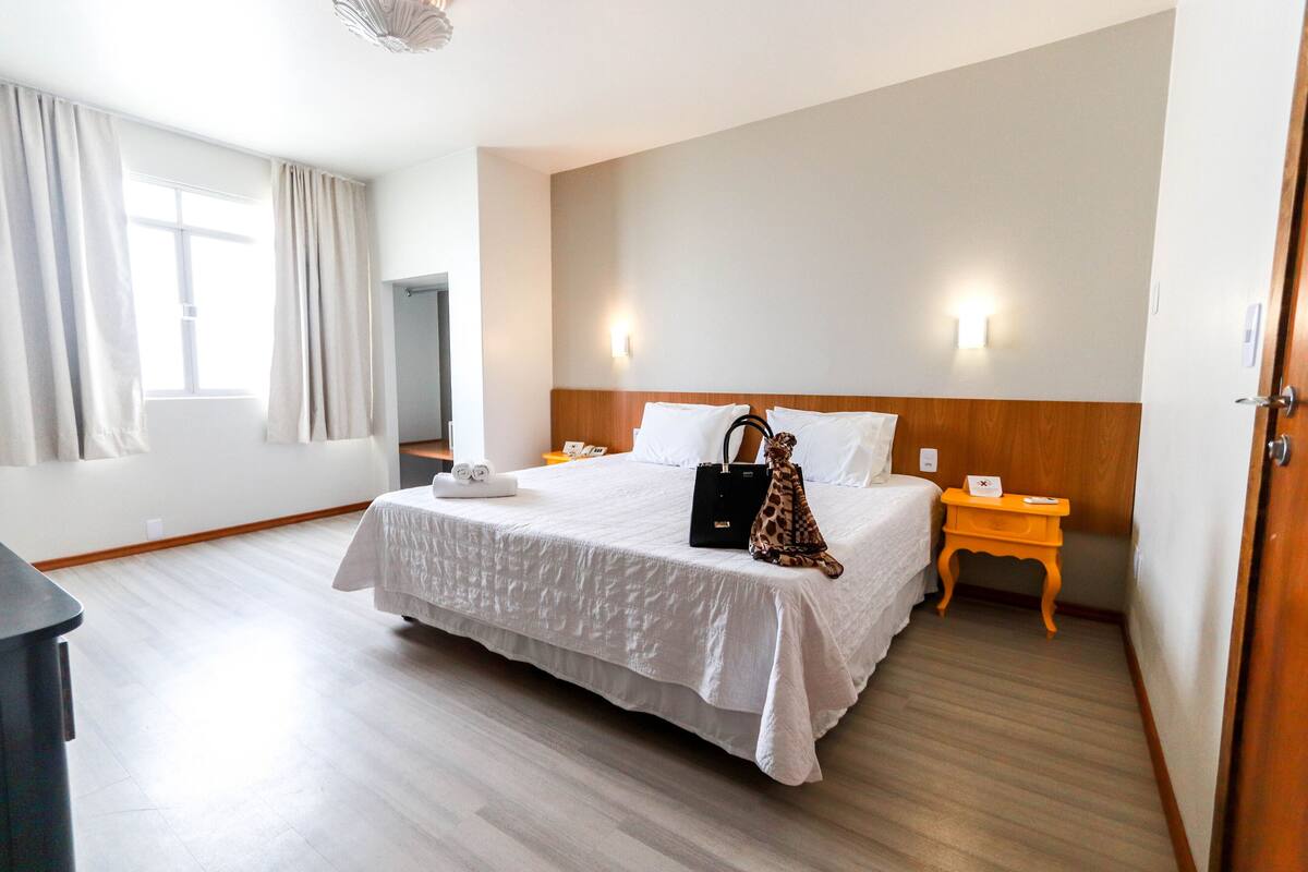 A hotel room with a large bed, a nightstand, a wooden headboard, a window with curtains, and a television.