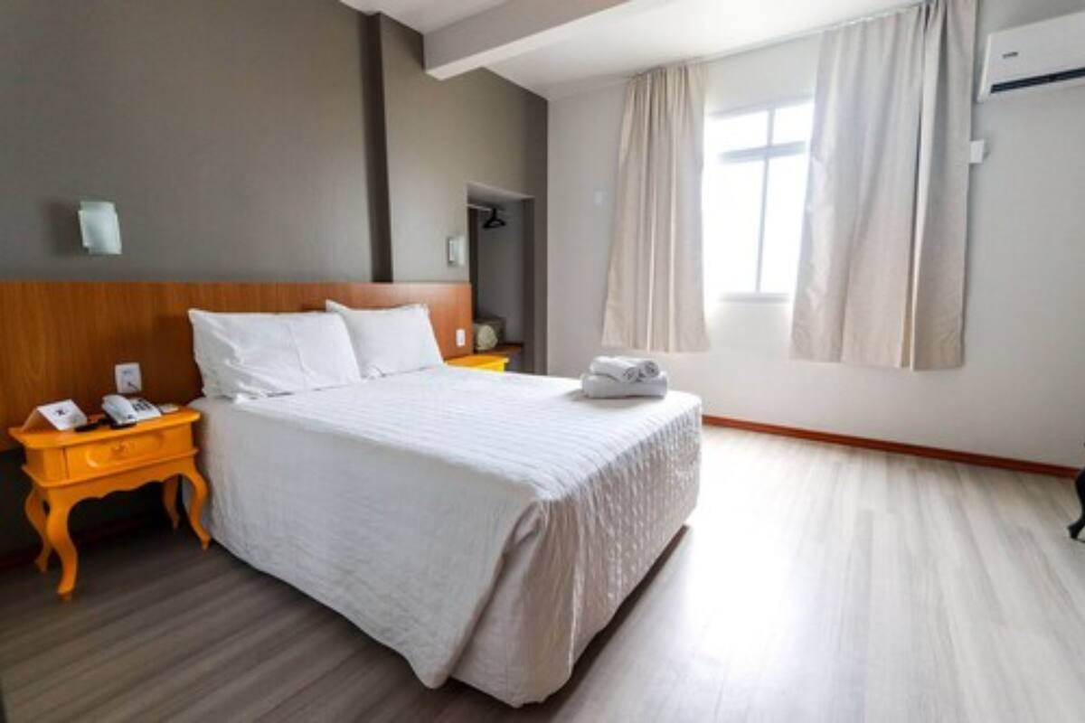 A hotel room with a bed, a nightstand with a phone, a window with curtains, and a wall-mounted air conditioner.