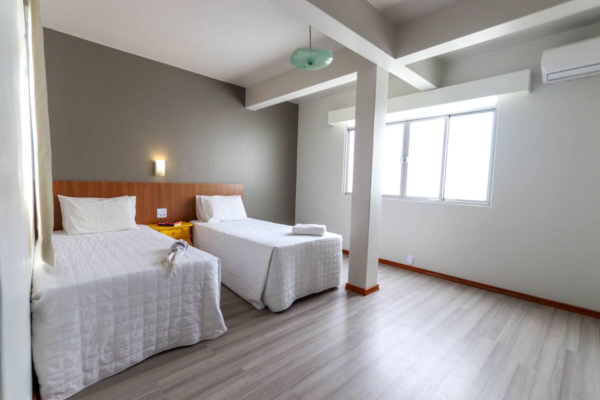 A hotel room with two beds, a wooden headboard, a small table, a nightstand, a wall-mounted lamp, and a large window.