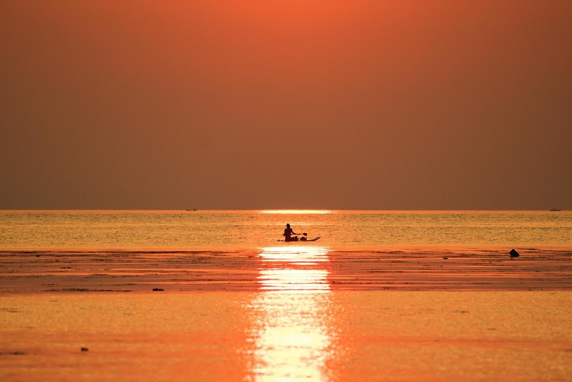 Een strandlandschap met iemand die op een boot zit, de zonsondergang weerspiegelt op het water.