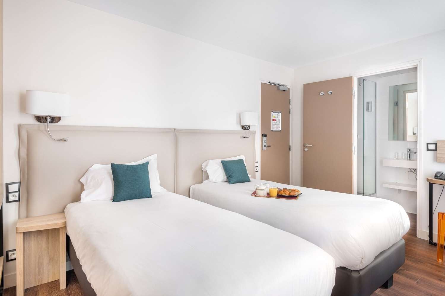A hotel room with two beds, a bedside table, a nightstand, a bathroom with a shower, and a small desk with a chair.