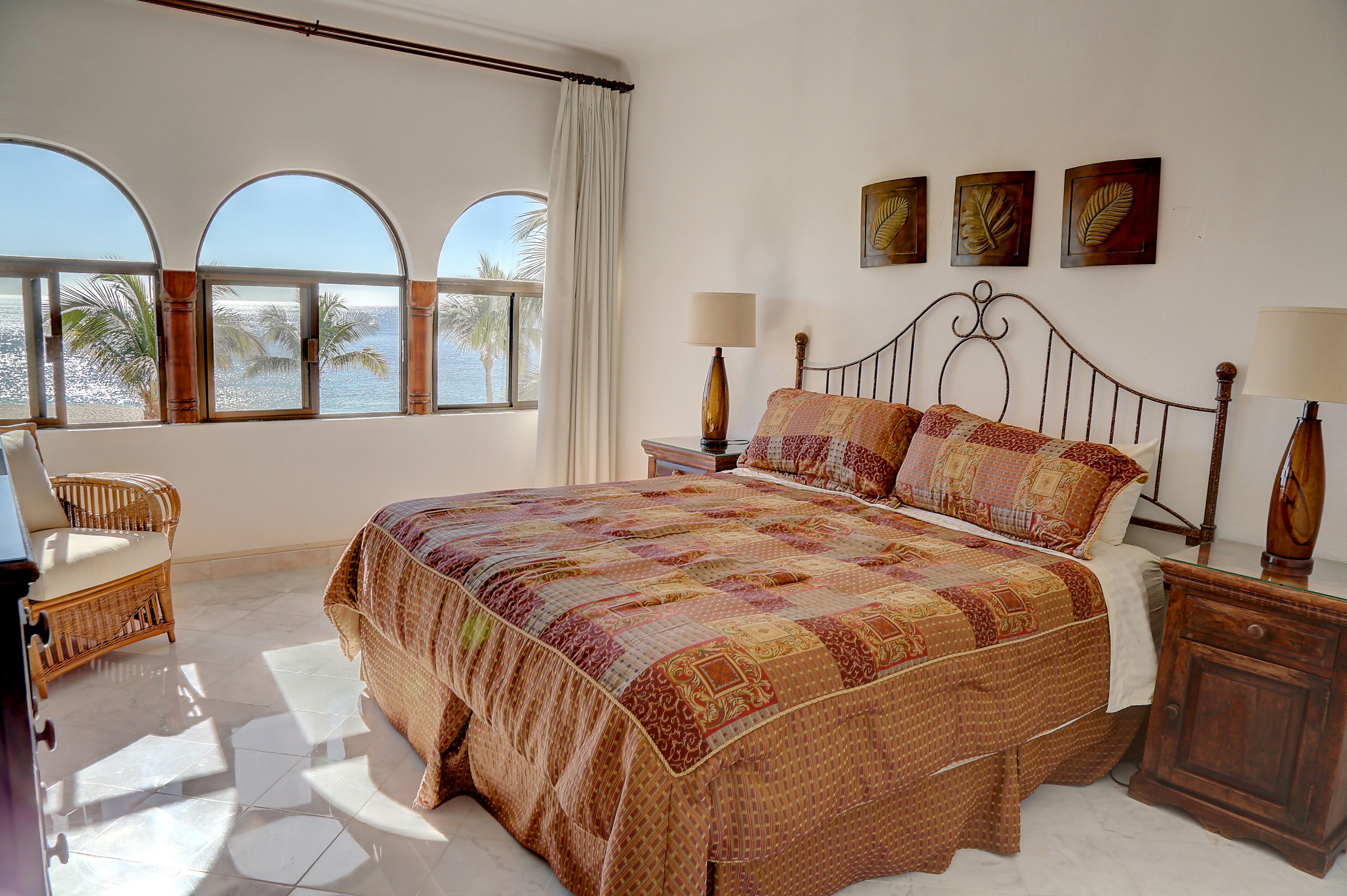 Un dormitorio con una cama grande, dos mesitas de noche, una silla y vistas al mar a través de ventanales arqueados.