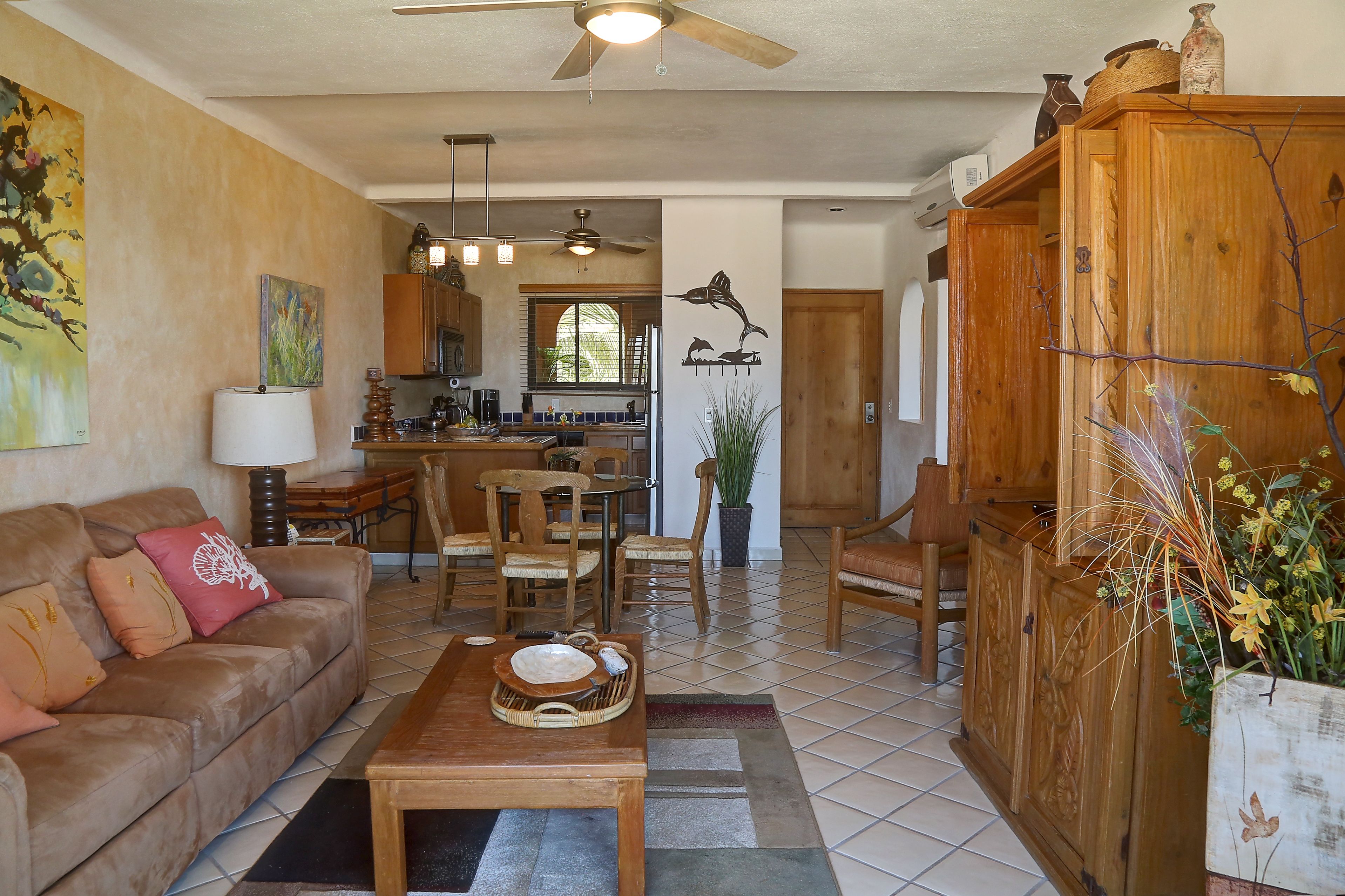 Amplia sala de estar con una mesa de centro de madera, un sofá y una zona de comedor con sillas y una mesa. Se observa una cocina al fondo, con armarios y una ventana.