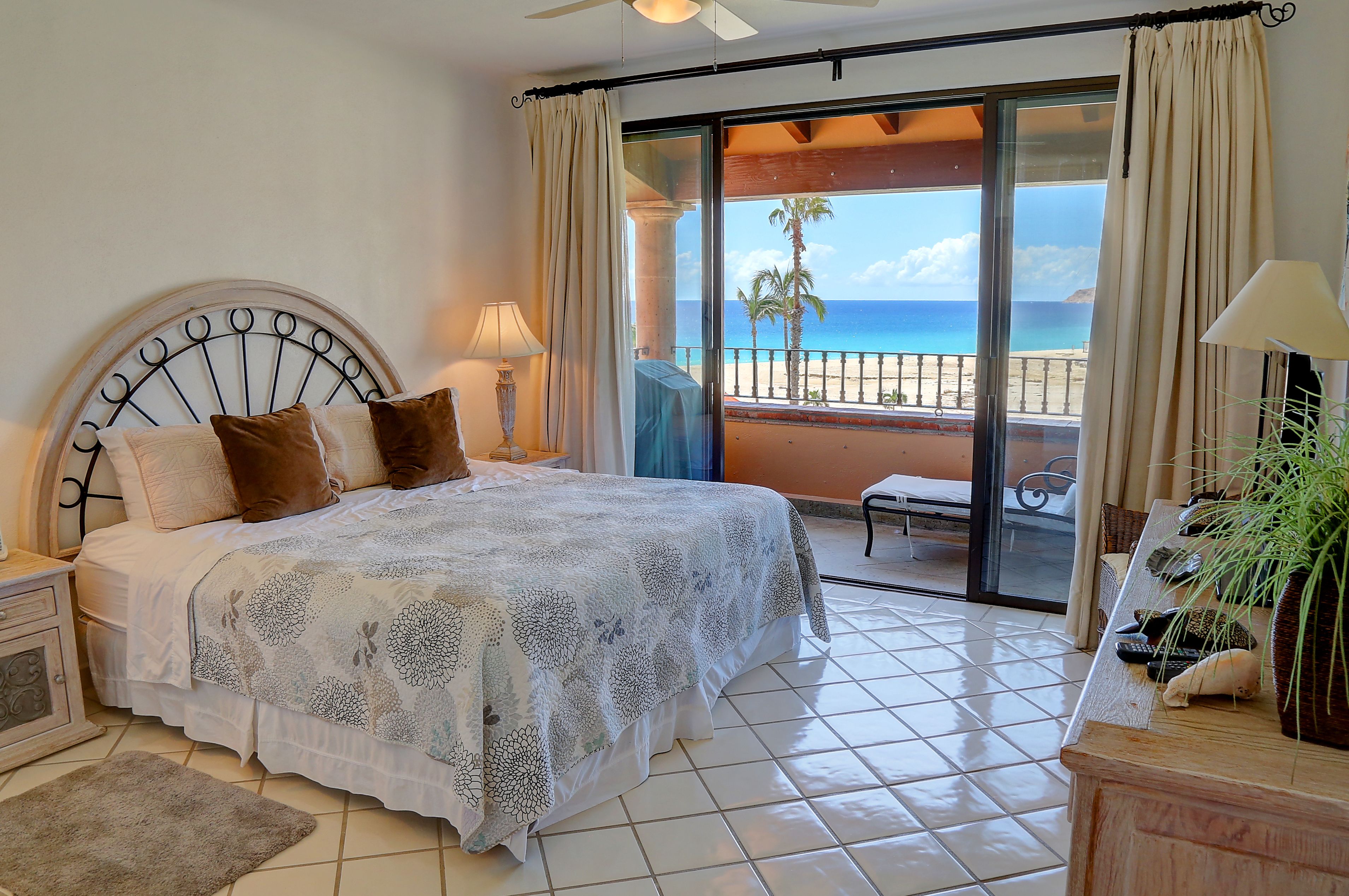 A bedroom with a bed, a nightstand, a lamp, a plant, and a view of the beach.