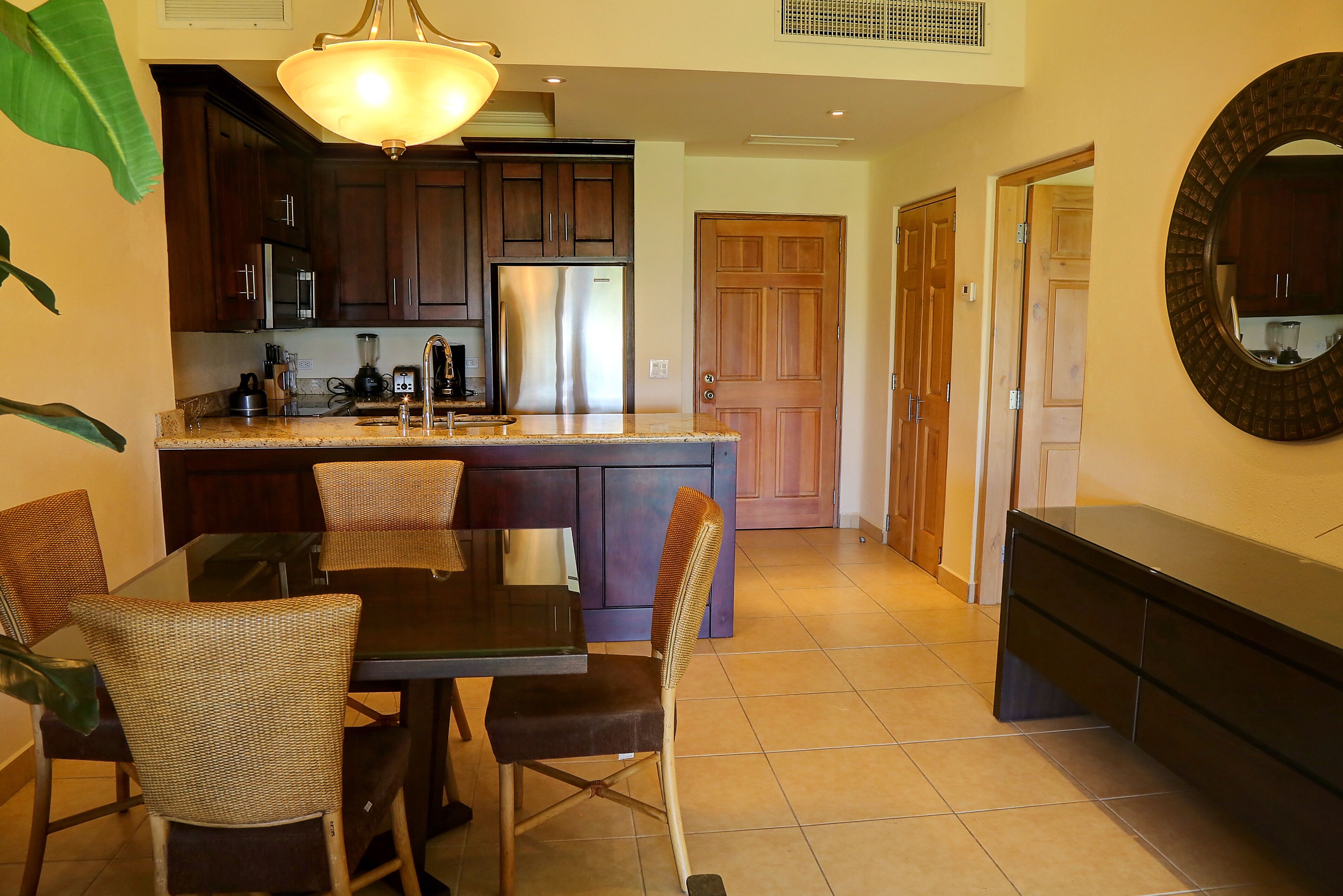 A modern kitchen with dark wooden cabinets, a stainless steel refrigerator, and a dining area with wicker chairs and a glass table.