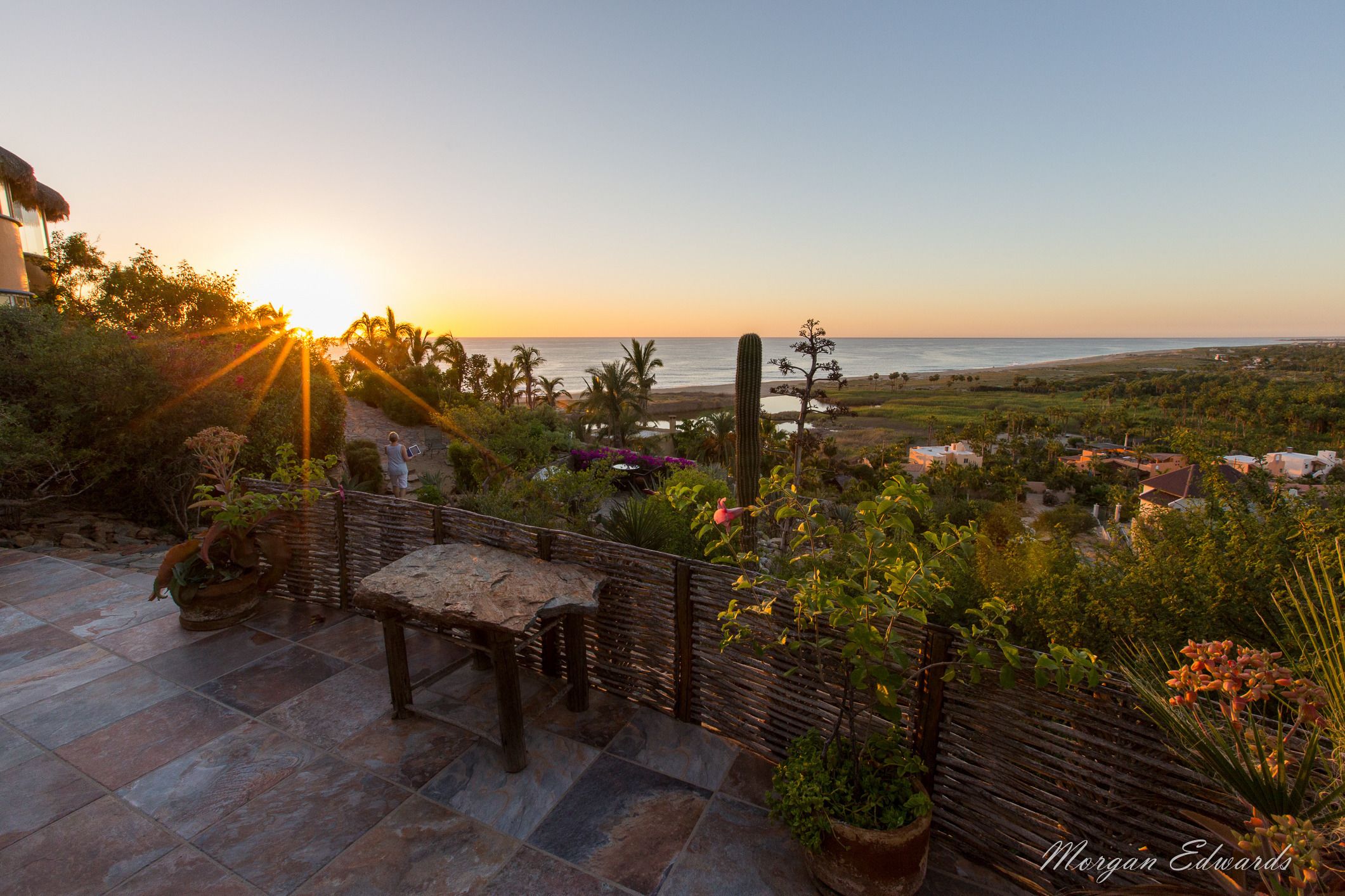 Une terrasse avec un sol en pierre, un banc en bois et une vue sur l’océan au coucher du soleil.
