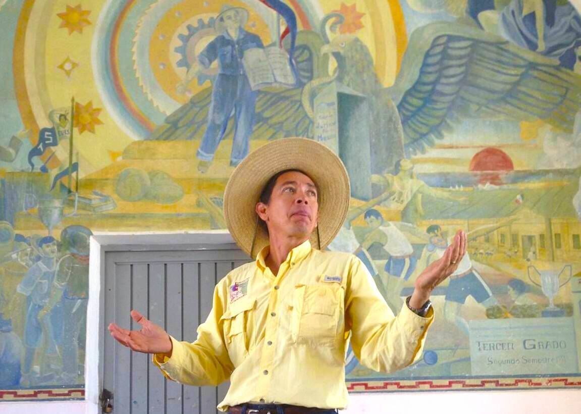 Una persona con camisa amarilla y sombrero de paja, de pie frente a un mural con varias figuras y un sol.