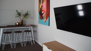 A modern office space with a large flat-screen TV mounted on the wall, a wooden desk with a vase of flowers, and three white bar stools. - City Quarters on Brewer (Perth)