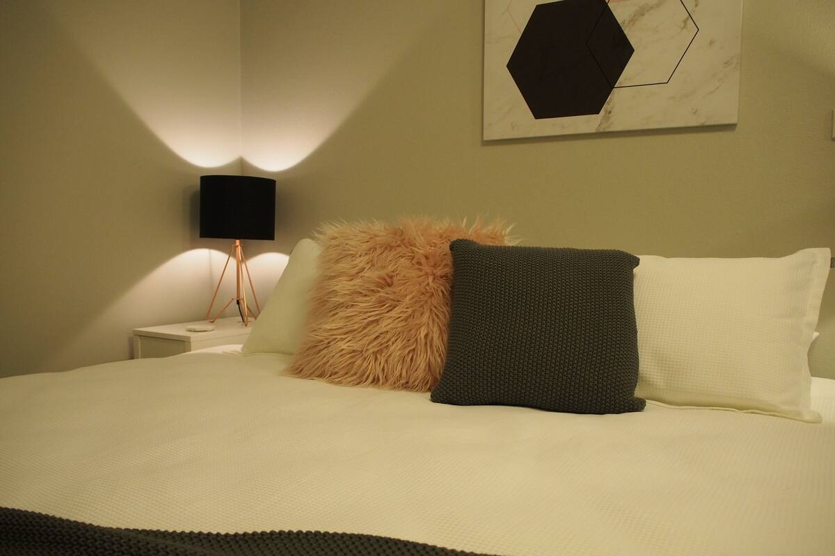 Una cama bien hecha con un almohadón mullido y uno oscuro, una lámpara de noche y una obra de arte colgada en la pared.