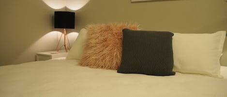 A neatly made bed with a fluffy pillow and a dark pillow, a bedside lamp, and a wall-mounted artwork.