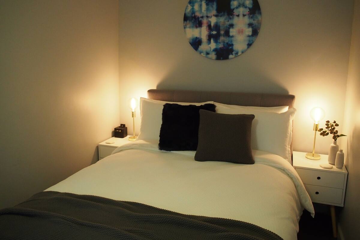 Una cama bien hecha con edredón blanco, dos almohadas y una manta oscura. Hay dos mesitas de noche, una con lámparas y un reloj. Encima de la cama hay una obra de arte redonda en la pared.