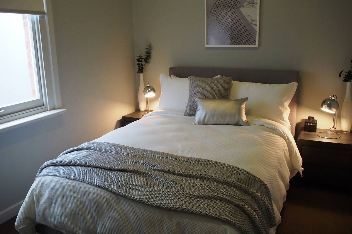 Una cama bien tendida con una colcha gris y blanca, dos lámparas de noche, un reloj y una obra de arte abstracta enmarcada en la pared.