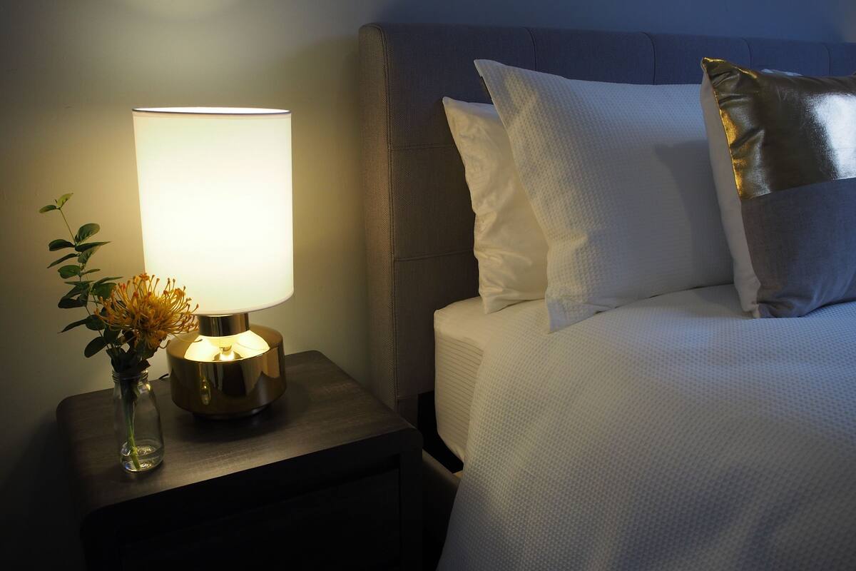 Una cama bien hecha con ropa blanca y una almohada dorada, una lámpara de noche con pantalla blanca y un pequeño jarrón con flores sobre una mesita de noche de madera.