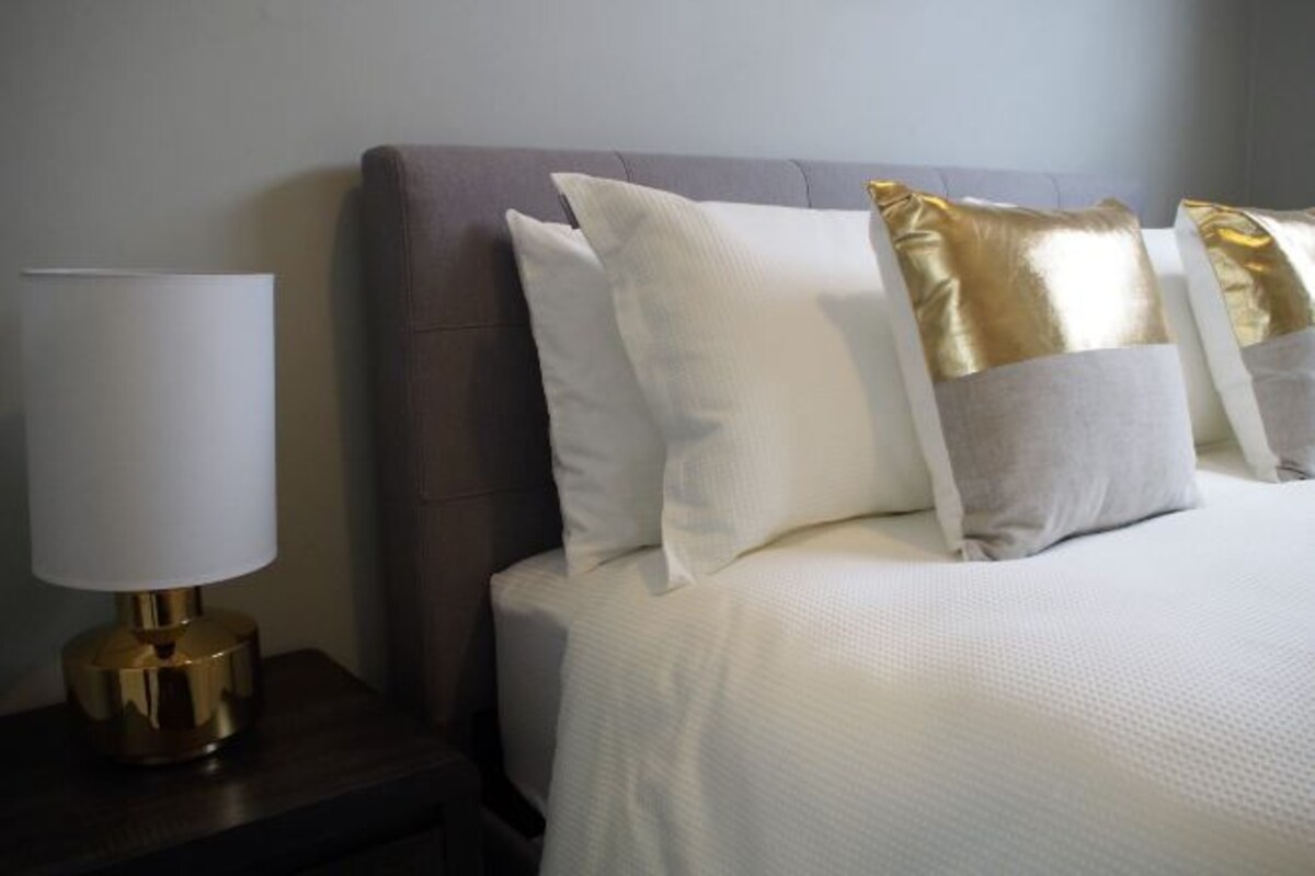 A neatly made bed with white bedding and gold and white pillows, a gold lamp on a wooden nightstand, and a dark-colored headboard.