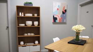 A dining area with a wooden table, white chairs, a bookshelf with dishes, and a vase of flowers. - City Quarters on Brewer (Perth)