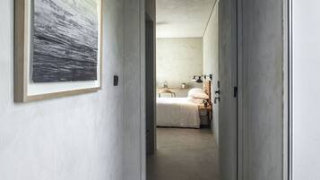Un couloir étroit mène à une chambre à coucher qui contient un lit, une table de chevet, une lampe et une œuvre d’art encadrée accrochée au mur.