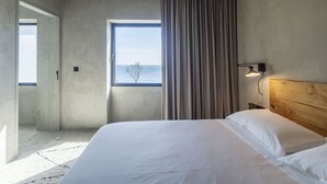 A modern hotel room with a large bed, a view of the sea through a window, and a wooden headboard.