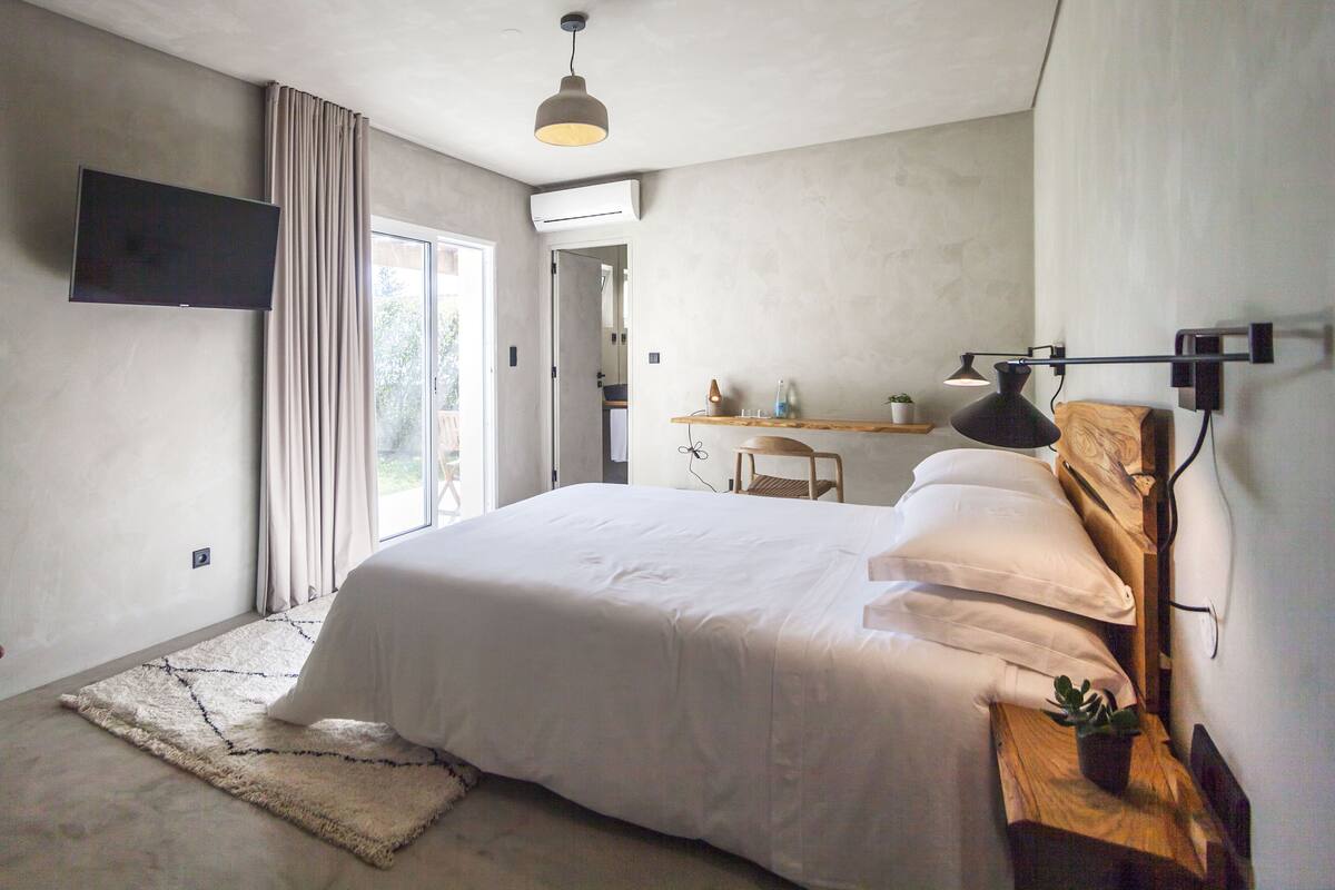 A modern bedroom with a large bed, a TV mounted on the wall, a desk with a chair, and a view of the outdoors.