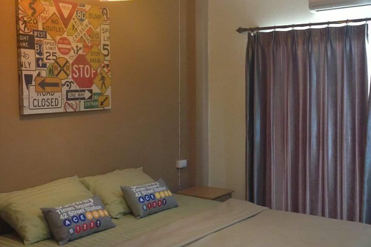 Una habitación de hotel con una cama, almohadas, un reloj de pared, un ventilador de techo y una ventana con cortinas.
