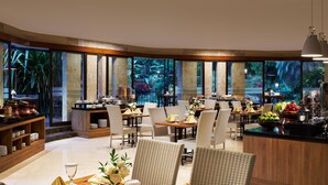 Een restaurant met meerdere eettafels en stoelen, een toonbank met een fruitschaal, en uitzicht op een tropische tuin door grote ramen.